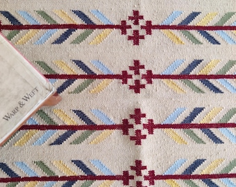 May include: A woven textile featuring a repeating pattern of horizontal maroon lines intersected by small maroon cross shapes. Diagonal segments in light blue, dark blue, yellow, and green flank the lines. A book corner with the text Warp & Weft is visible.