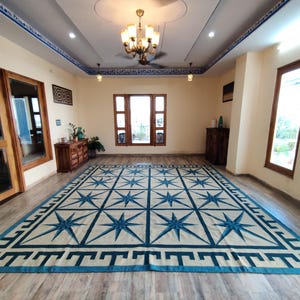 May include: A large rectangular rug with a cream-colored field and a repeating pattern of blue eight-pointed stars. The rug has a blue border with a geometric design. The room has wooden doors and windows, and a chandelier.
