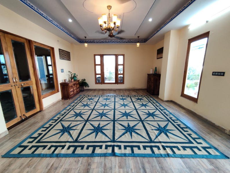 May include: A large rectangular rug with a cream-colored field and a repeating pattern of blue eight-pointed stars. The rug has a blue border with a geometric design. The room has wooden doors and windows, and a chandelier.