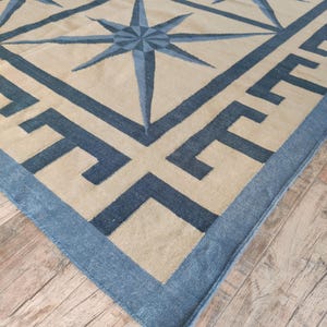 May include: A large, rectangular rug with a geometric design. The rug features a beige background with a pattern of blue compass rose stars and a blue border with a Greek key design. The rug is made of a woven material.