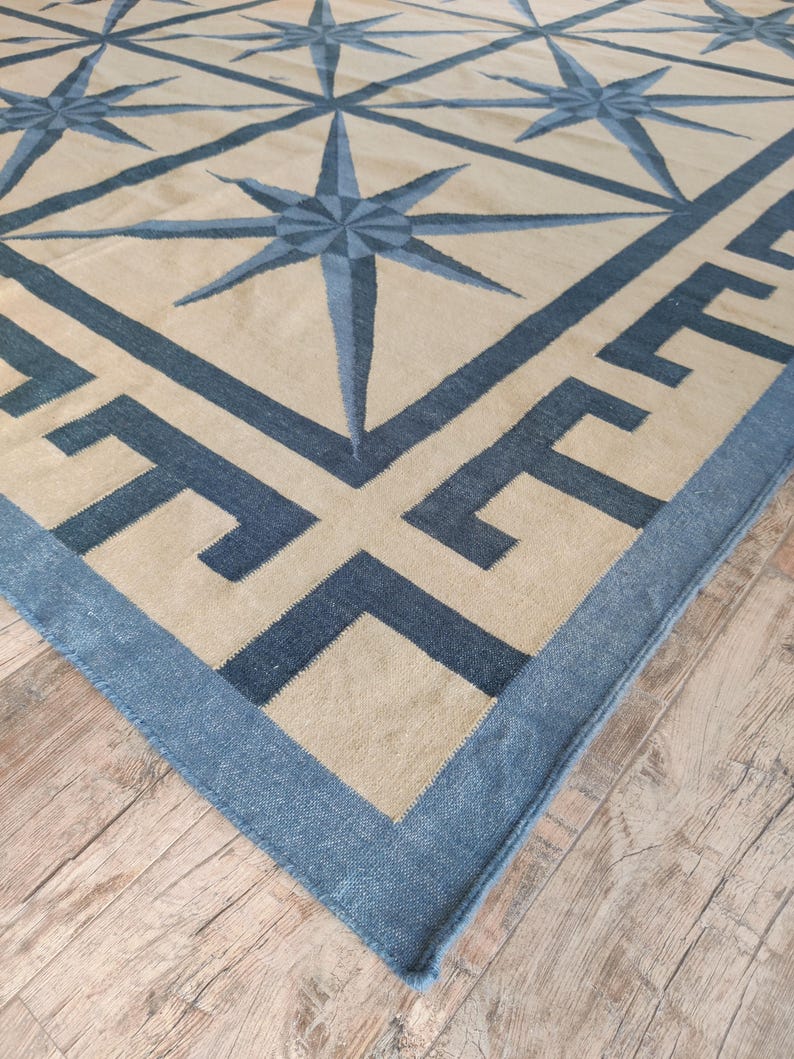 May include: A large, rectangular rug with a geometric design. The rug features a beige background with a pattern of blue compass rose stars and a blue border with a Greek key design. The rug is made of a woven material.