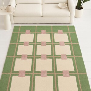 May include: A rectangular green rug with a geometric pattern. The design features beige squares, pink rectangles, and tan lines. The rug is placed on a white floor, with a white sofa in the background.