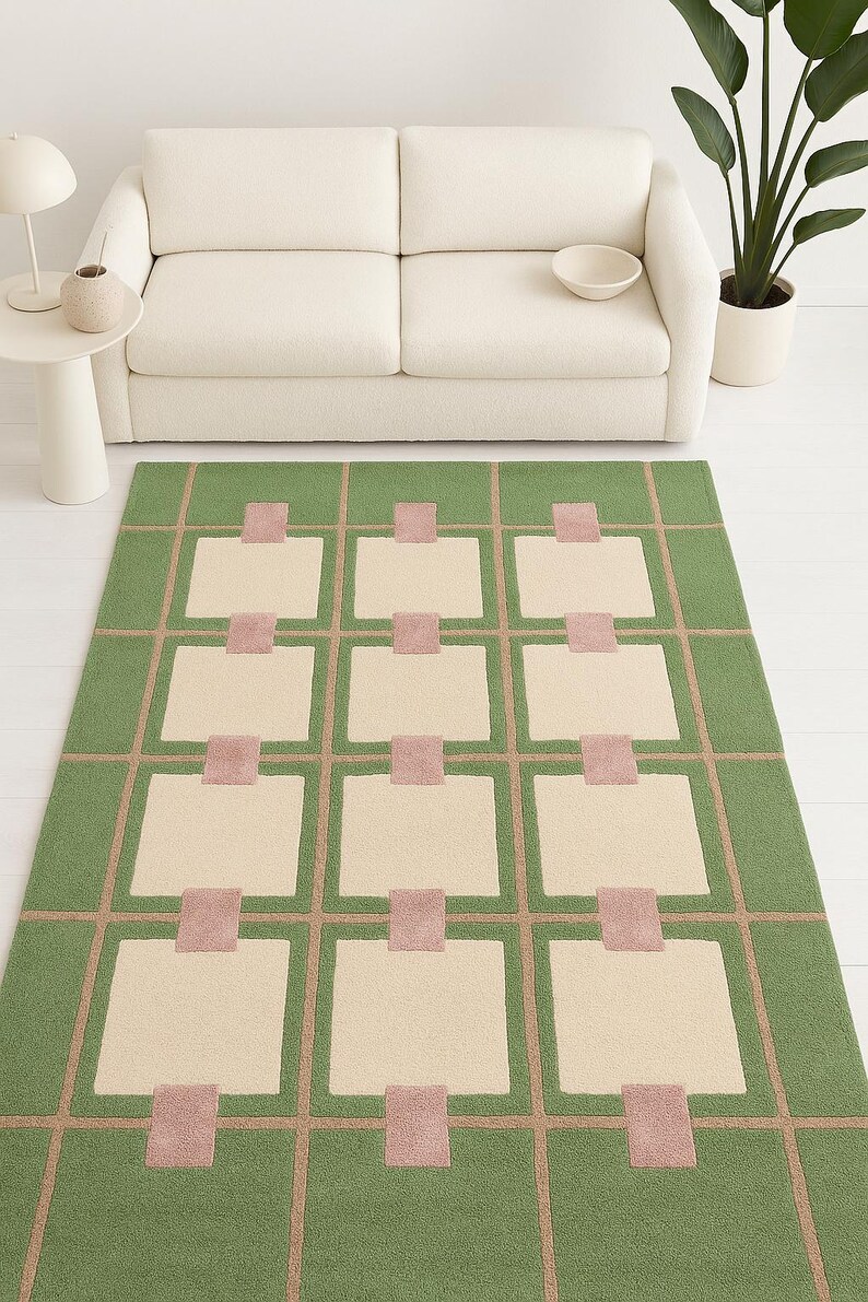 May include: A rectangular green rug with a geometric pattern. The design features beige squares, pink rectangles, and tan lines. The rug is placed on a white floor, with a white sofa in the background.