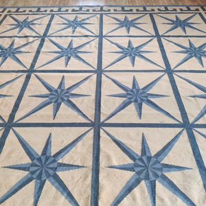 May include: Large beige and blue area rug with a repeating pattern of eight-pointed stars. The stars are in a lighter blue shade and are framed by a darker blue grid. The rug has a border with a Greek key design.