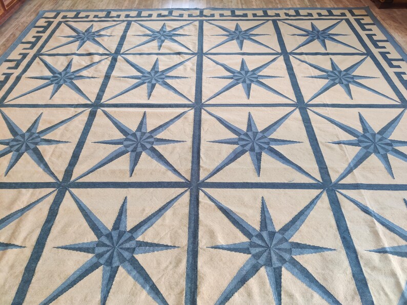 May include: Large beige and blue area rug with a repeating pattern of eight-pointed stars. The stars are in a lighter blue shade and are framed by a darker blue grid. The rug has a border with a Greek key design.