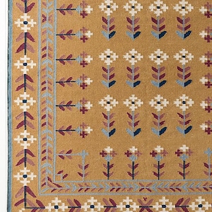 Może przedstawiać: Dywan w kolorze musztardowym z powtarzającym się wzorem kwiatowym w kolorze białym, bordowym i niebieskim. Dywan ma obramowanie z geometrycznymi i kwiatowymi wzorami. Frędzle dywanu są widoczne wzdłuż dolnej krawędzi.