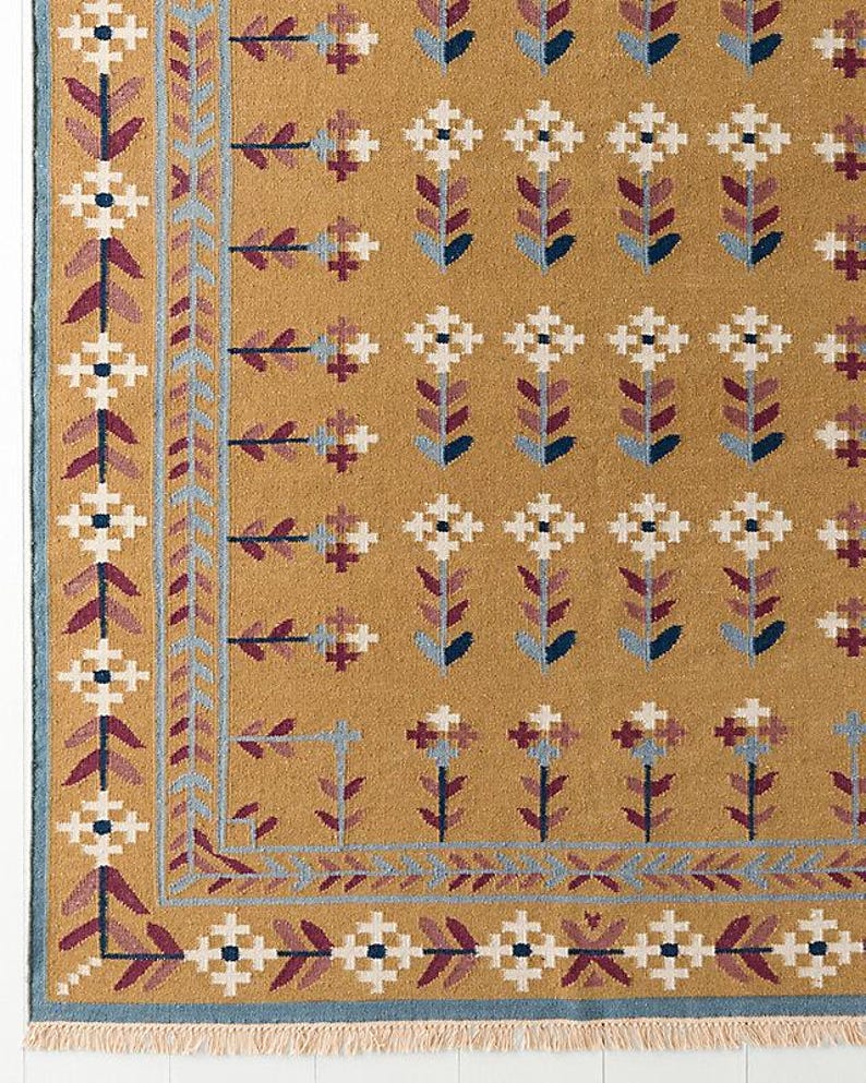 May include: Mustard yellow area rug with a repeating floral pattern in white, burgundy, and blue. The rug has a border with geometric and floral designs. The rug's fringe is visible along the bottom edge.