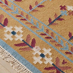 May include: Close-up of a tan rug with a geometric pattern. The rug features blue, white, and burgundy floral and leaf designs. The rug has a fringe border.