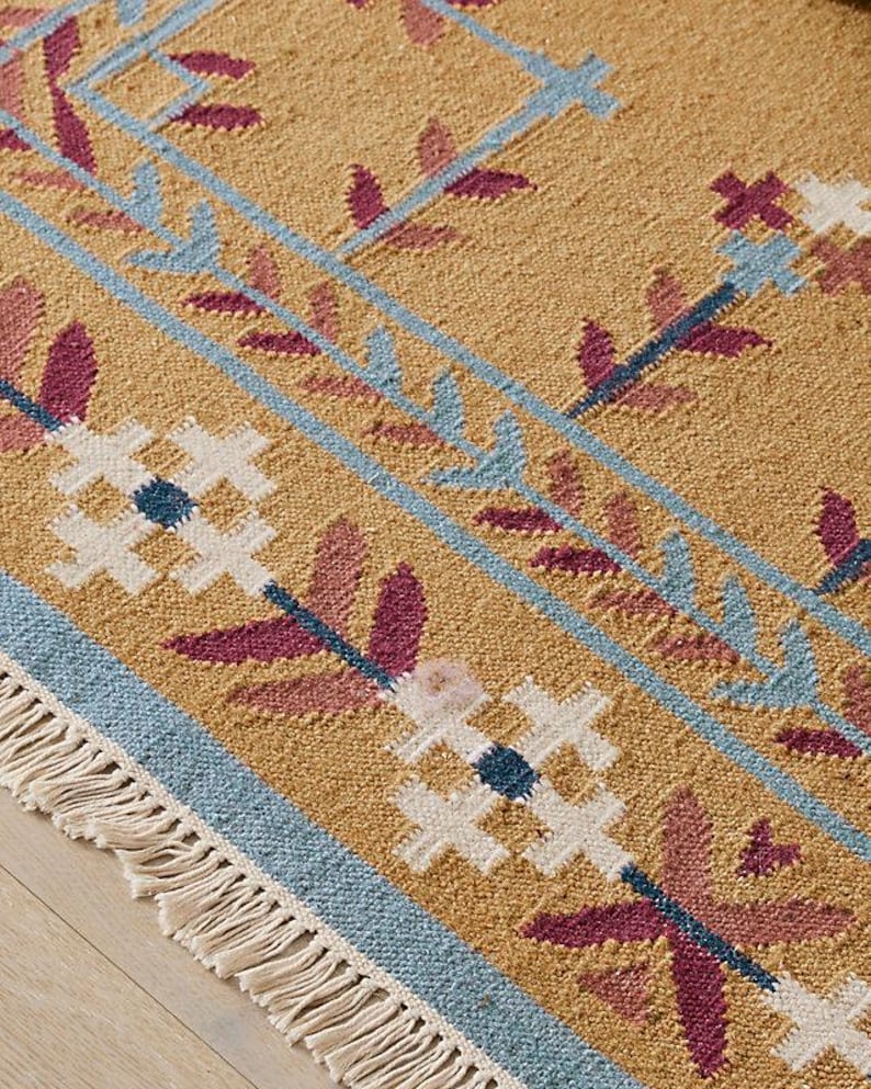 May include: Close-up of a tan rug with a geometric pattern. The rug features blue, white, and burgundy floral and leaf designs. The rug has a fringe border.