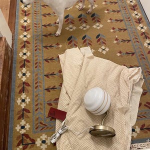 May include: A white and gold globe lamp with a round base and a white cord sits on a beige fabric with small polka dots. A red tag is attached to the fabric. A dog with white and brown fur stands on a patterned rug.