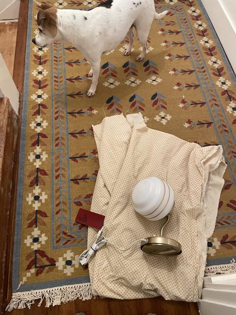 May include: A white and gold globe lamp with a round base and a white cord sits on a beige fabric with small polka dots. A red tag is attached to the fabric. A dog with white and brown fur stands on a patterned rug.
