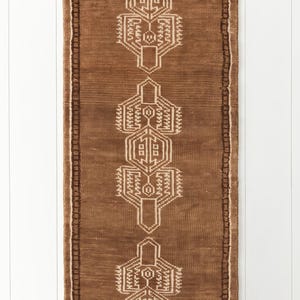 May include: A rectangular runner rug in a deep brown hue featuring a series of three symmetrical, cream-colored geometric motifs aligned vertically. The rug is framed by a dark brown border with a repeating pattern and finished with fringe at both ends.
