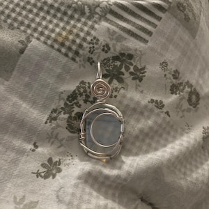 Pode incluir: Um pingente envolto em arame com uma pedra azul-clara lisa de formato oval. O fio de metal prateado é enrolado ao redor do perímetro da pedra e forma um laço circular no topo para uma corrente, criando uma moldura decorativa segura.