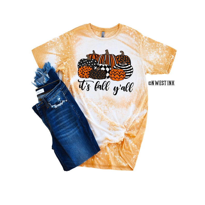 May include: A bleached yellow t-shirt with a graphic of a pile of orange, black, and white pumpkins with the text "it's fall y'all". The shirt is paired with blue jeans.