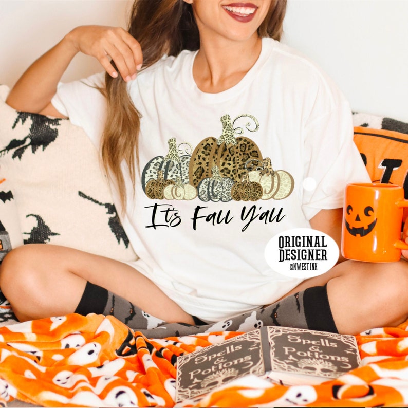 May include: White t-shirt with a graphic of a stack of pumpkins with leopard print and gold accents. The text "It's Fall Y'all" is printed below the pumpkins. The text "Original Designer @NWESTINK" is printed in the bottom right corner of the shirt.