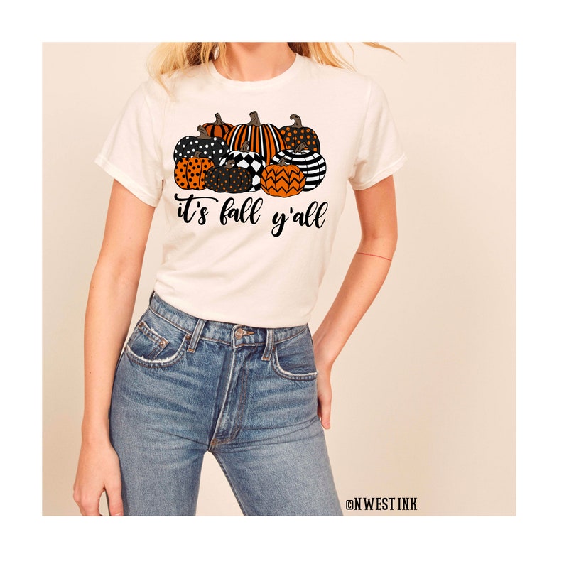May include: White t-shirt with a graphic of orange, black, and white pumpkins and the text "it's fall y'all".