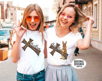 May include: Two white t-shirts with a leopard print cross and the text "He is Risen". One shirt features a brown bunny rabbit standing on the cross.