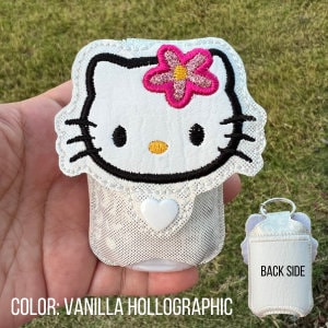 May include: A white and cream-colored Hello Kitty-themed hand sanitizer holder with a pink flower and black accents. The holder has a heart-shaped clasp and is labeled "COLOR: VANILLA HOLLOGRAPHIC". The back side is also shown.