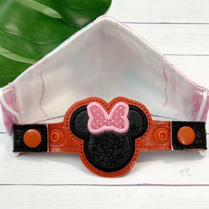 May include: A pink and white fabric face mask with a black and pink Minnie Mouse character patch attached to a brown strap with two orange buttons.