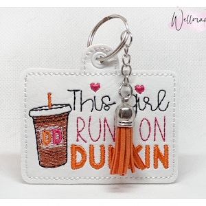 Könnte beinhalten: Weißer Leder-Schlüsselanhänger mit einem braunen und orangefarbenen gestickten Dunkin' Donuts Kaffeebecher und dem Text "This girl runs on Dunkin'" mit einem rosa Herz über dem Text und einem rosa Herz über dem Wort "girl". Der Schlüsselanhänger hat einen silbernen Quasten.