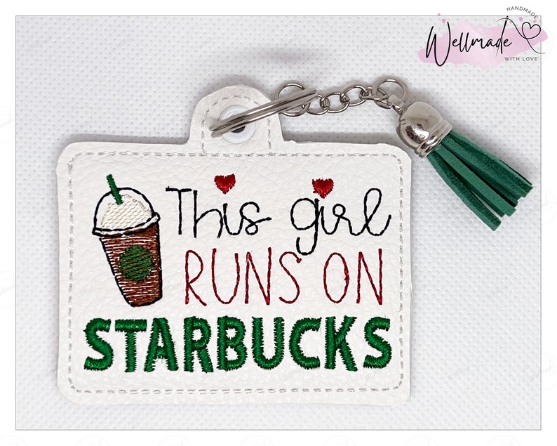 May include: White leather keychain with a green tassel and embroidered text that reads "This girl runs on Starbucks". The keychain features a red heart and a cartoon image of a Starbucks coffee cup.