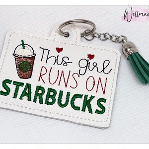 May include: White leather keychain with a green and brown embroidered image of a Starbucks coffee cup and the text "This girl runs on Starbucks". The keychain has a green tassel.