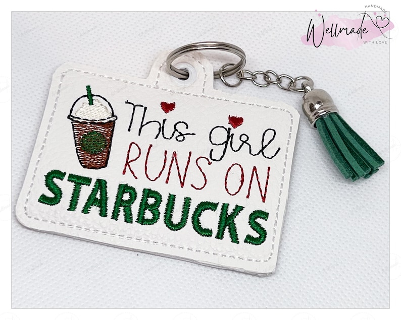 May include: White leather keychain with a green and brown embroidered image of a Starbucks coffee cup and the text "This girl runs on Starbucks". The keychain has a green tassel.