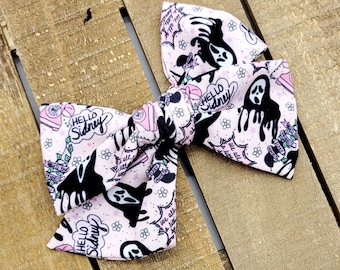 Puede incluir: Un lazo para el pelo de tela con fondo rosa claro y un patrón repetitivo de figuras negras fantasmales, teléfonos de disco rosas, pequeñas flores y burbujas de texto que dicen Hello Sidney y What's Your Favorite Scary Movie.