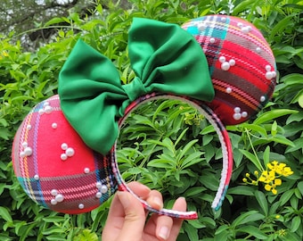 May include: A festive headband with two round ears covered in red plaid fabric, adorned with white pearl accents. A large green bow sits between the ears. The headband is held against a backdrop of green foliage.
