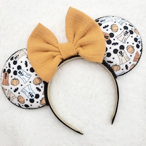 May include: A pair of black and white mouse ears with a Halloween theme. The ears feature a pattern of ghosts, pumpkins, and Mickey Mouse heads. A large mustard yellow bow is attached to the headband.