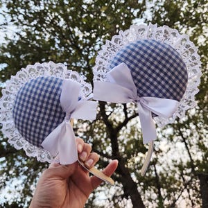 Puede incluir: Dos diademas a cuadros azules y blancos con ribete de encaje blanco y lazos de satén blanco. Las diademas se sostienen sobre un fondo de follaje verde.