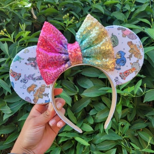 May include: A pair of white mouse ears with a colourful sequined bow. The ears feature a white background with colourful cartoon characters from the film Inside Out.