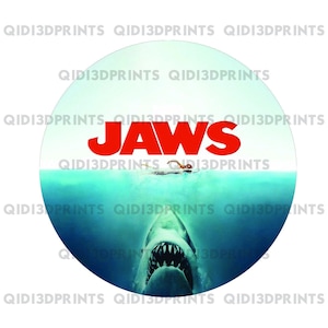 Op de afbeelding: Een ronde sticker met een blauwe en groene oceaan scène. De tekst "JAWS" staat in rode letters boven het water. Een haai zwemt met open bek omhoog vanaf de bodem van het water. Een persoon zwemt in het water boven de haai.