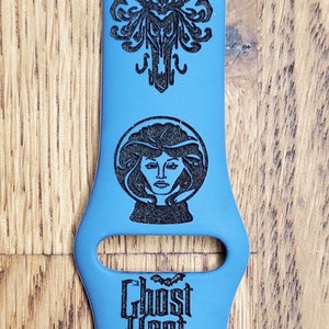 May include: A blue silicone Apple Watch band with a black engraved design featuring the Haunted Mansion, a ghost, a woman in a crystal ball, and the text "Ghost Host".