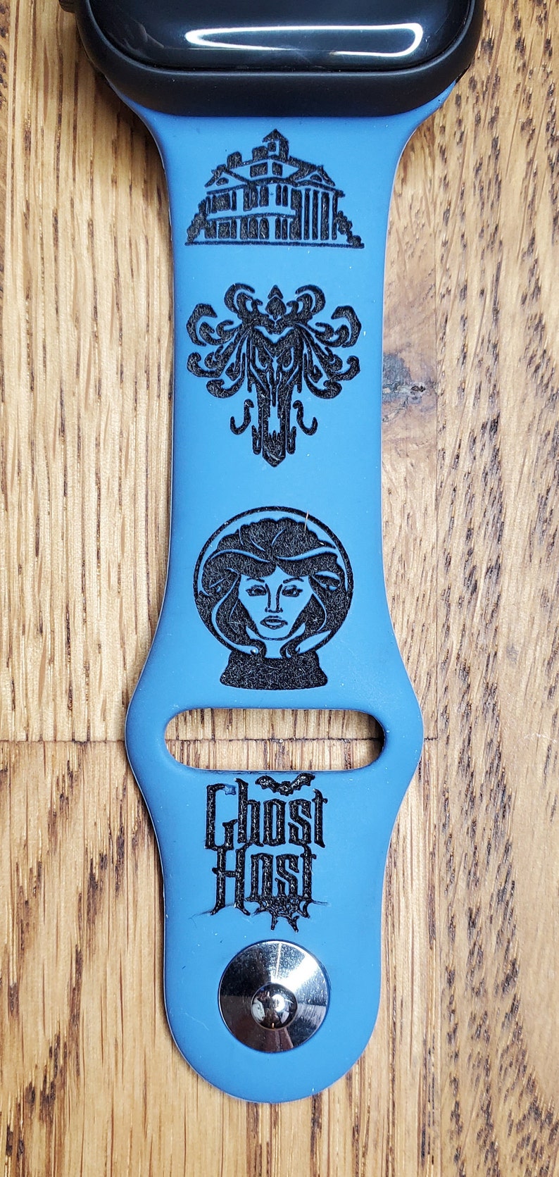 May include: A blue silicone Apple Watch band with a black engraved design featuring the Haunted Mansion, a ghost, a woman in a crystal ball, and the text "Ghost Host".