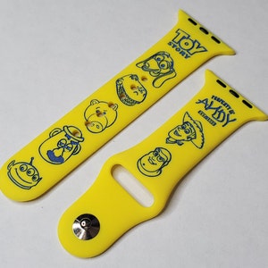 May include: Yellow silicone Apple Watch band with blue Toy Story characters, including Woody, Buzz Lightyear, and Jessie. The band says "Property of Andy" and "Toy Story" in blue text.