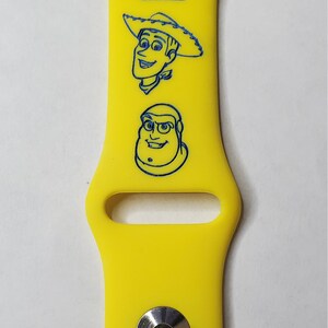 May include: Yellow silicone watch band with blue outline images of Woody and Buzz Lightyear from Toy Story. The band has the text "PROPERTY OF ANDY EST. 1995" printed on it.