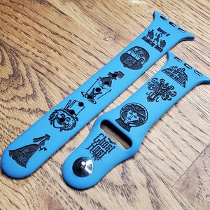 May include: Two blue Apple Watch bands with black engraved designs featuring characters and text from the Haunted Mansion attraction at Disneyland. The bands feature the text "Ghost Host" and "Beware of Hitchhiking Ghosts".