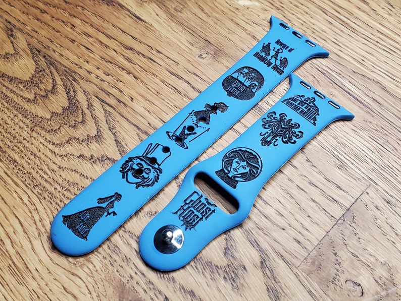 May include: Two blue Apple Watch bands with black engraved designs featuring characters and text from the Haunted Mansion attraction at Disneyland. The bands feature the text "Ghost Host" and "Beware of Hitchhiking Ghosts".