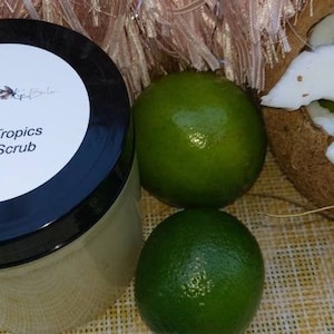 May include: A glass jar of sugar scrub with a black lid and a white label that reads "In The Tropics Sugar Scrub". The jar is surrounded by limes and a coconut shell.