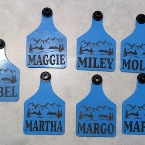 May include: Seven blue plastic livestock identification tags with black mountain and lake scenes and black text. The tags read "Mabel", "Maggie", "Miley", "Molly", "Martha", "Margo", and "Maple".