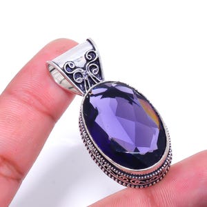 May include: A silver pendant with an oval, faceted, deep purple gemstone. The pendant has an ornate silver bail with decorative swirls. The gemstone is set in a silver bezel with a detailed border. The pendant is held by a person's fingers.