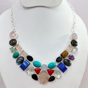 以下が含まれることがあります： 様々な色の宝石が付いたシルバーチェーンのネックレス。ピンク、青、緑、黒の石など、さまざまな形と色があります。ネックレスは白いマネキンに飾られています。