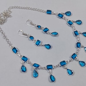 Könnte beinhalten: Ein silbernes Halsketten- und Ohrring-Set mit blauen tropfenförmigen und quadratischen Edelsteinen. Die Halskette hat ein Kettenglied-Design und die Ohrringe sind im Hänge-Stil.