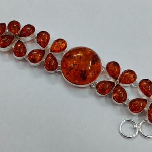 May include: A silver bracelet featuring amber stones. The bracelet has a central large, round amber stone, surrounded by smaller, teardrop-shaped amber stones. The bracelet has a toggle clasp.