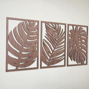 May include: Three wooden wall decor pieces featuring cutout tropical leaf designs. The leaves are in shades of brown and are arranged in a horizontal row.