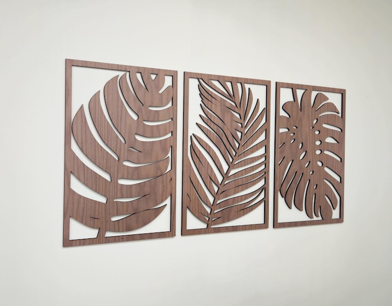 May include: Three wooden wall decor pieces featuring cutout tropical leaf designs. The leaves are in shades of brown and are arranged in a horizontal row.