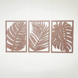 May include: Three wooden wall decor pieces featuring cutout tropical leaf designs. The pieces are arranged in a row, with a palm leaf in the center and monstera leaves on either side.
