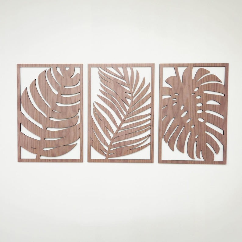 May include: Three wooden wall decor pieces featuring cutout tropical leaf designs. The pieces are arranged in a row, with a palm leaf in the center and monstera leaves on either side.