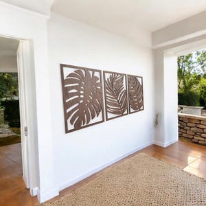 May include: Three wooden wall decor panels featuring cutout tropical leaf designs. The panels are brown and are arranged in a row on a white wall.